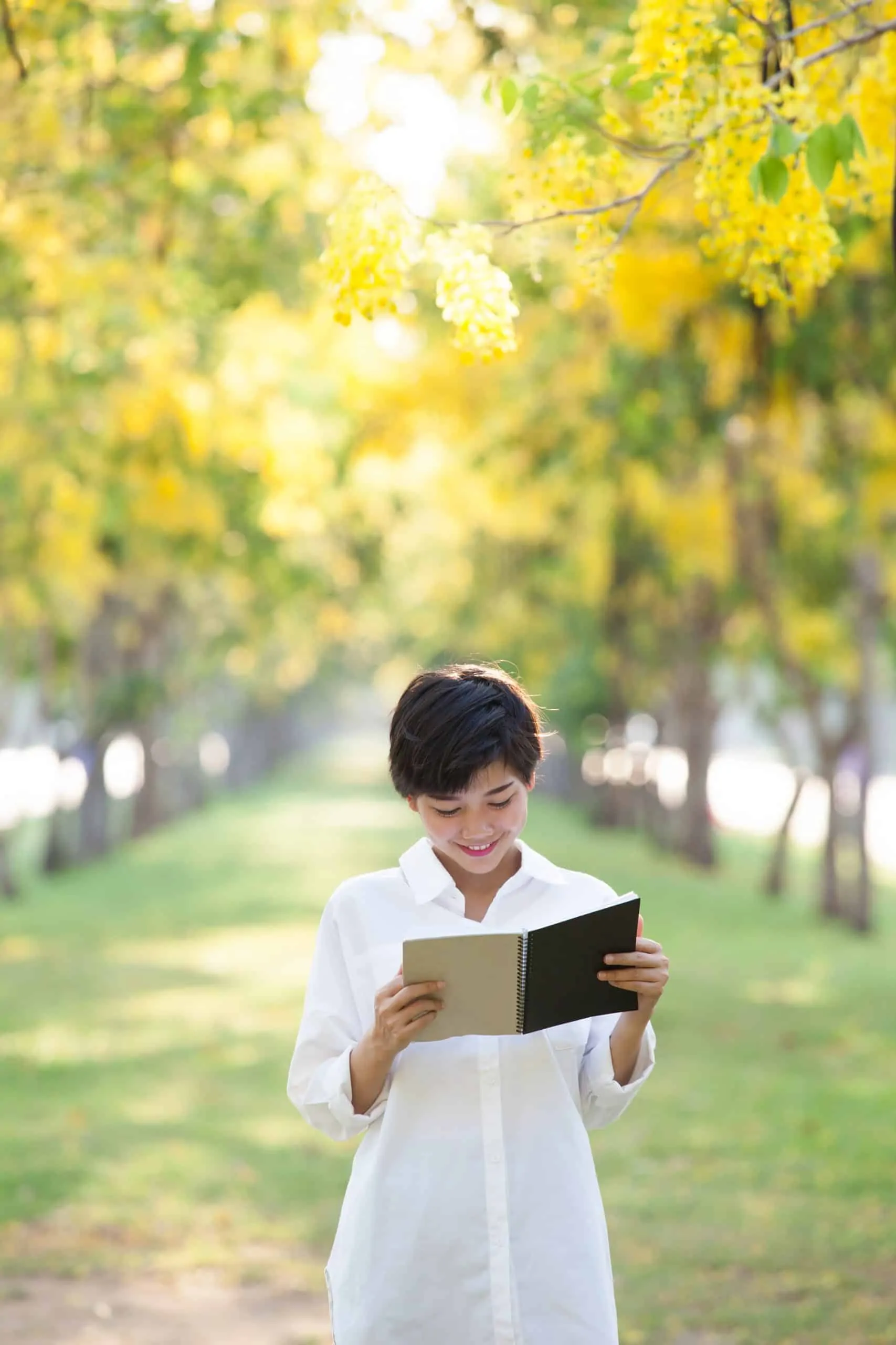 A young Asian man reading from a book from within a corridor of trees.