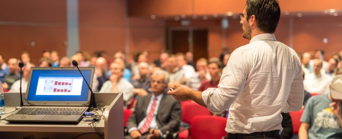A speaker at a conference in the foreground his seated audience in the background.